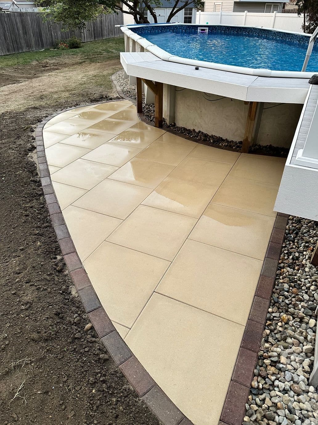 Newly paved poolside patio with beige tiles and stone edging, surrounded by a garden area.